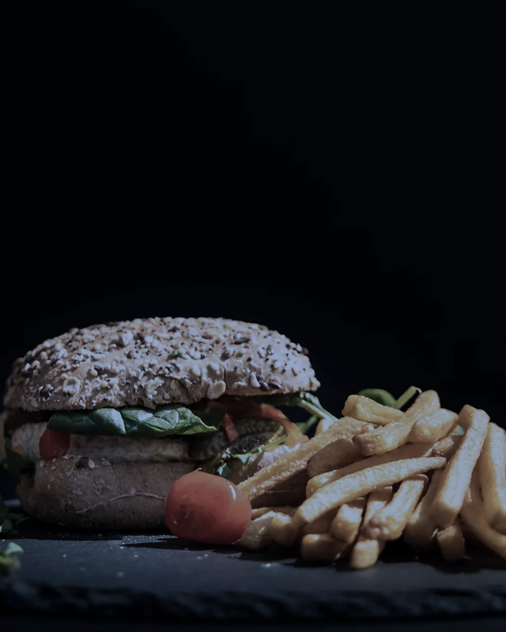 Hambúrguer com batata frita e tomate sobre uma pedra escura.