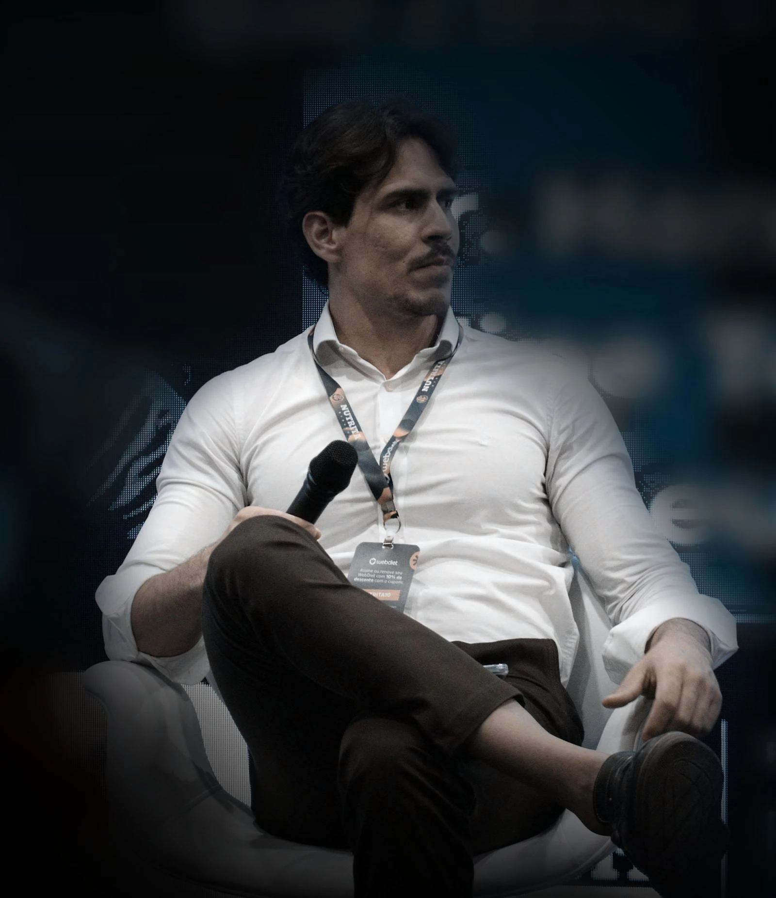 Matheus Dias Mendes sentado no palco de um evento, de camisa clara e crachá, com microfone na mão.