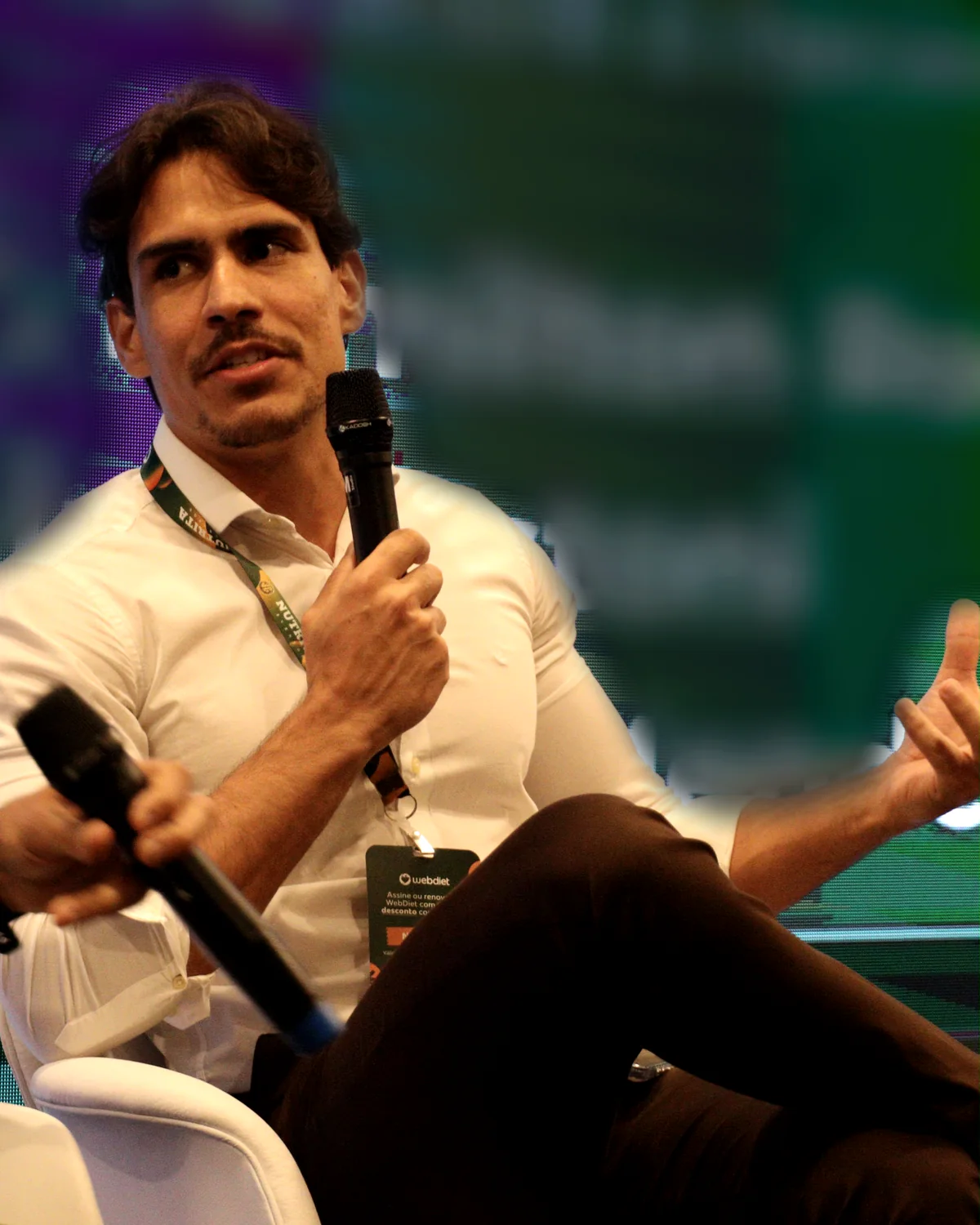 Matheus Dias Mendes falando ao microfone num painel de evento, de camisa branca e crachá.